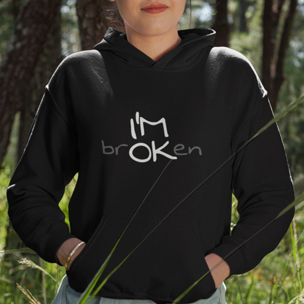 I'M brOKen Premium Sober Apparel worn by a woman in a black hoodie, featuring a hidden "I'M OK" message that symbolizes resilience, recovery, and hope through life's challenges.