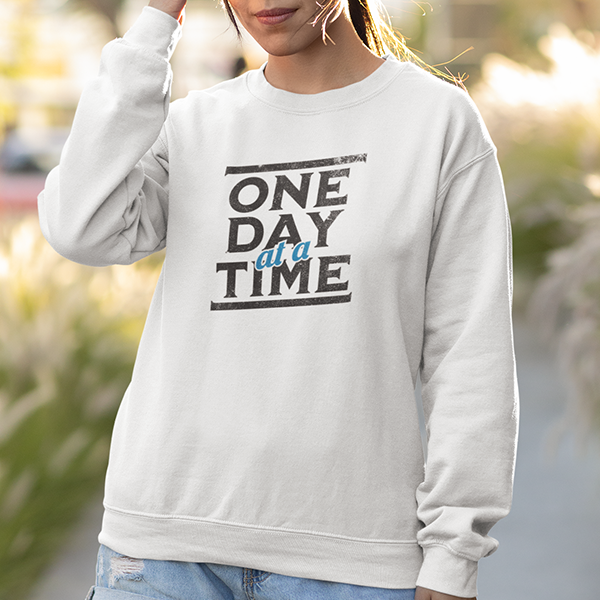 One Day At A Time Premium Sober Apparel worn by a woman in a white sweatshirt, highlighting a classic recovery principle that encourages living in the moment and moving forward with purpose.