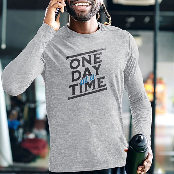 One Day At A Time Premium Sober Apparel worn by a smiling man in a sports gray long-sleeve shirt, representing the strength found in taking recovery one step at a time.